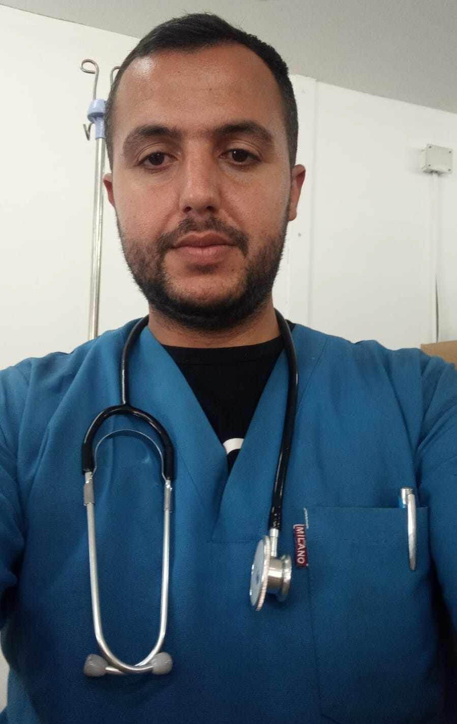 A photograph of Raed wearing blue scrubs. He has a stethoscope hanging from his neck.